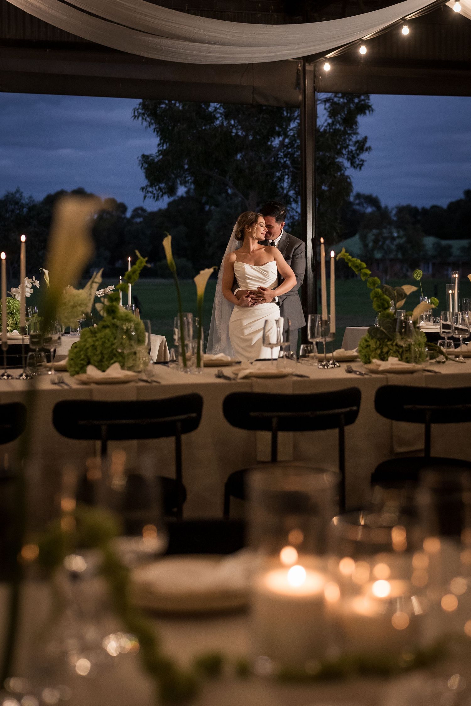 Bride and groom embrace at candlelit outdoor wedding reception at dusk under string lights.