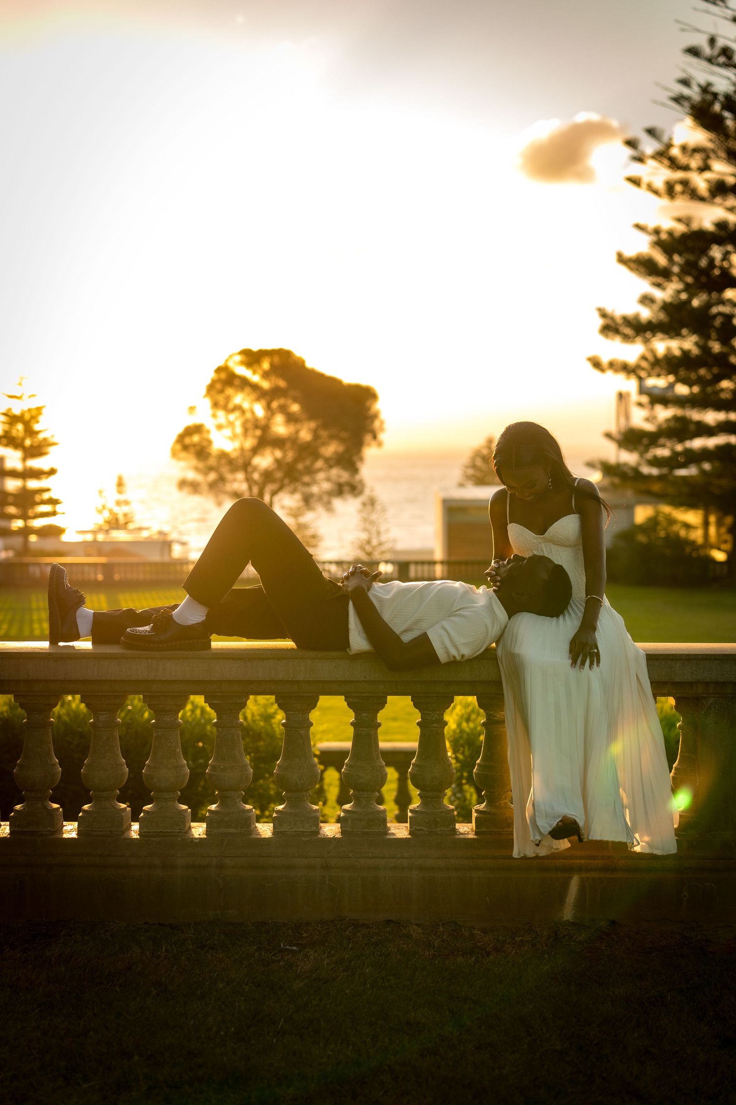 Couple in a romantic sunset portrait, a man lying on a stone balustrade as a woman in a white dress leans over him.