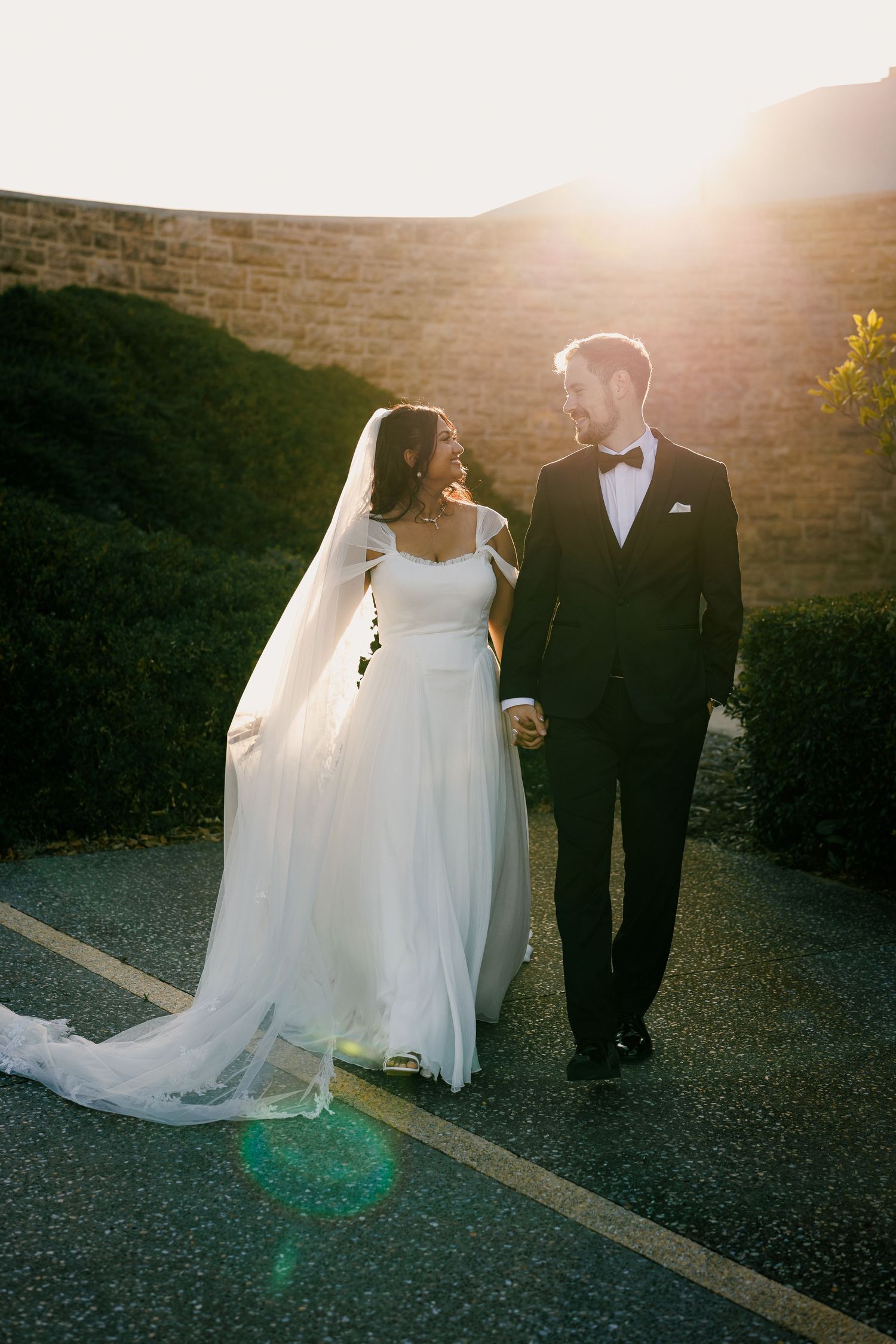 A bride and groom walk hand-in-hand at sunset, with golden backlight illuminating her flowing white gown and veil.