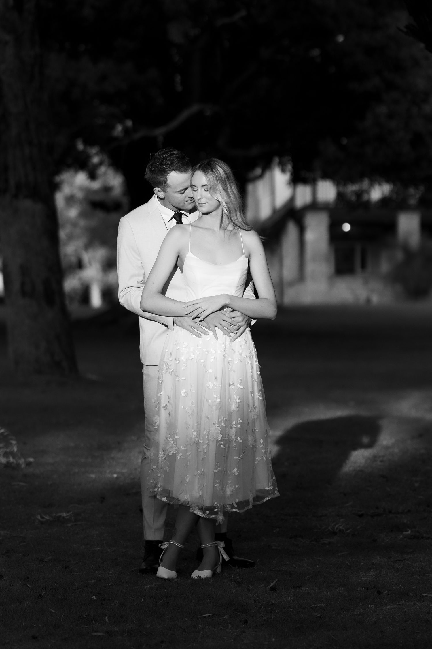 Black and white photo of a couple embracing outdoors at night, groom in white suit kissing bride in floral dress.