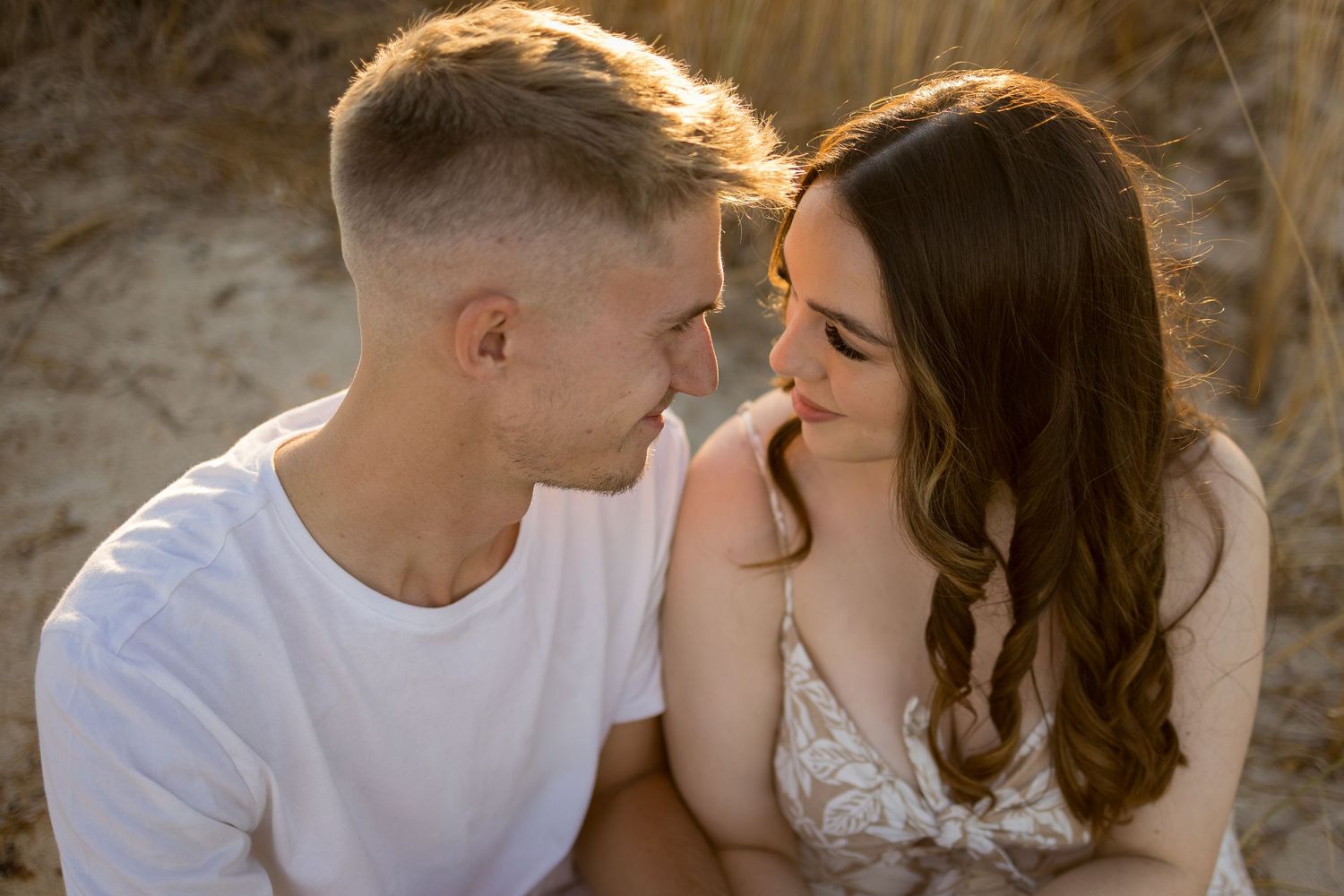 Romantic couple sharing an intimate moment outdoors, sitting close together in warm golden sunlight near sandy dunes.