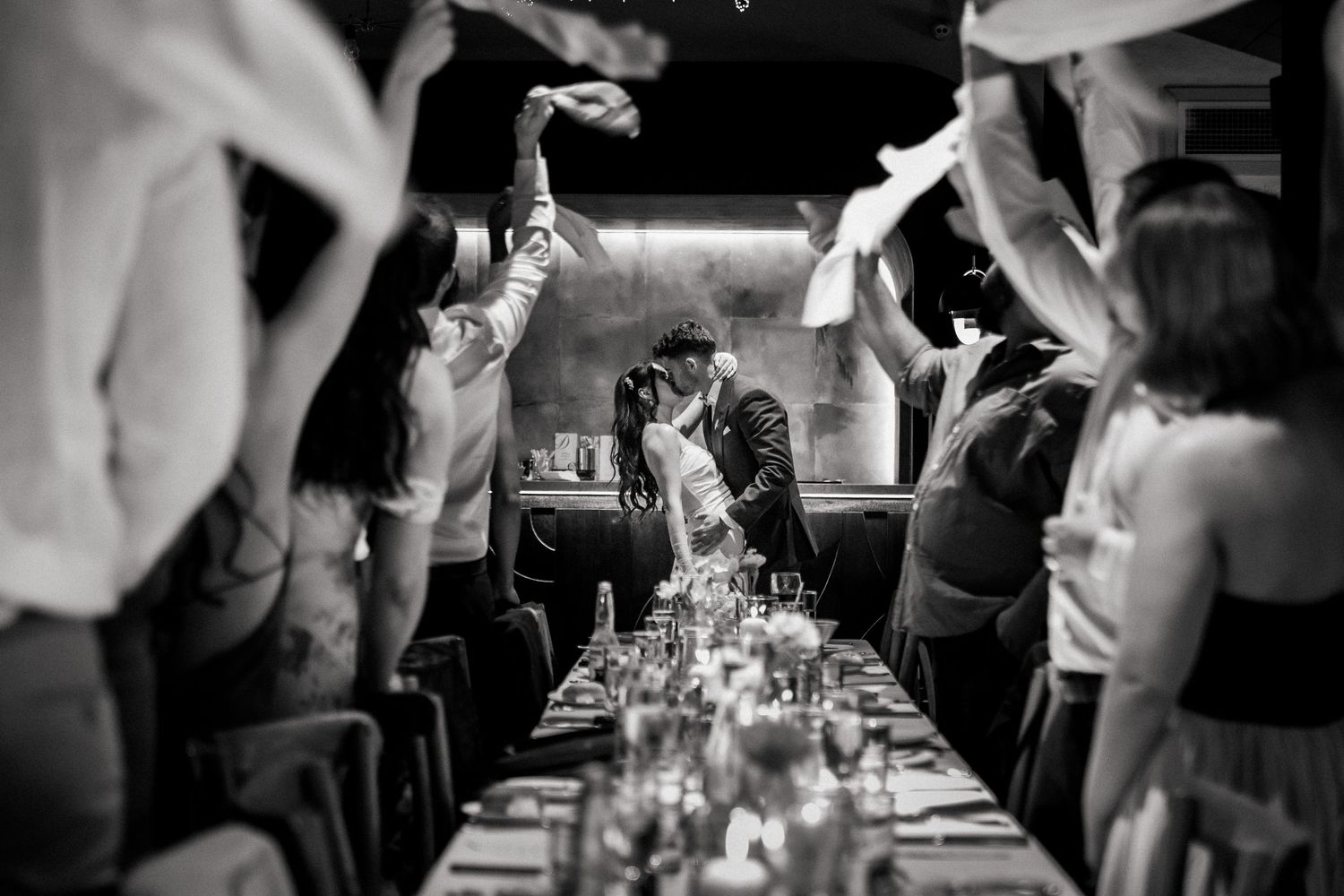 Couple shares a kiss at wedding reception as guests wave napkins in celebration.