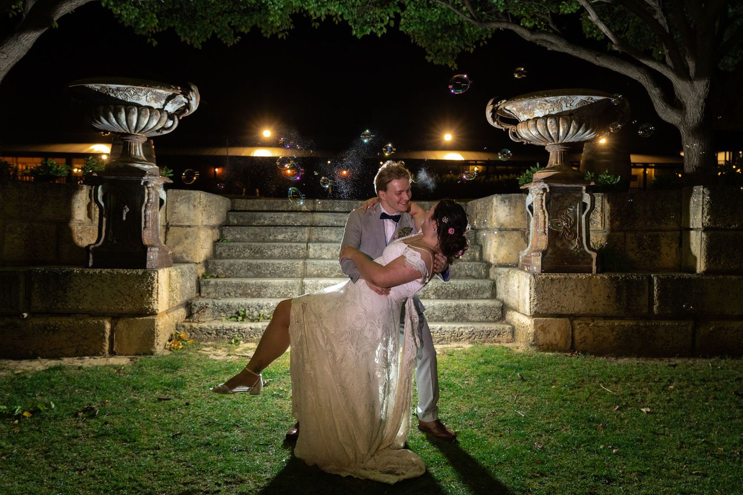 Bride and groom share romantic dip pose at night in front of elegant stone staircase with decorative urns.