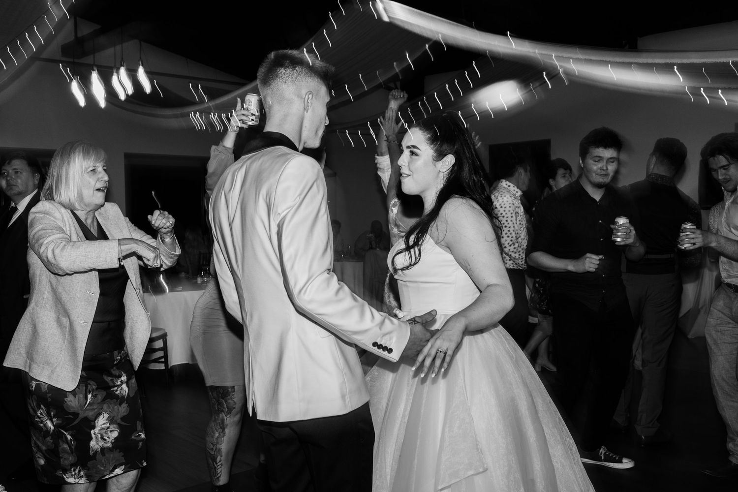 Black and white photo of a couple sharing their first dance at a wedding reception with guests celebrating in background.