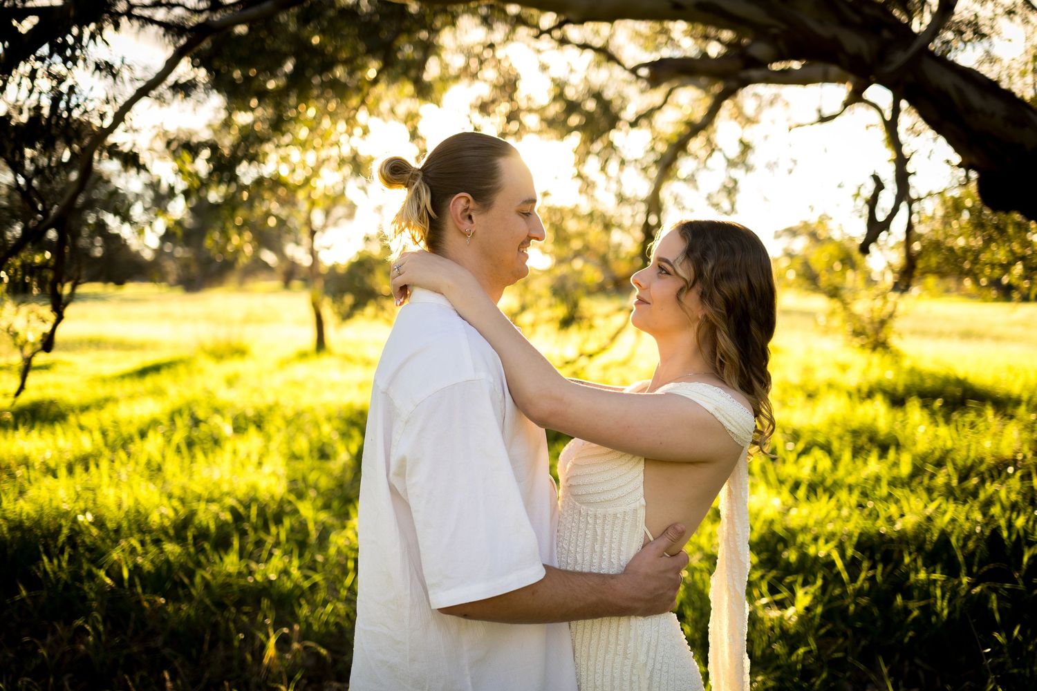 Couple embracing at golden hour in a sunlit green meadow with trees, wearing white outfits in a romantic outdoor setting.