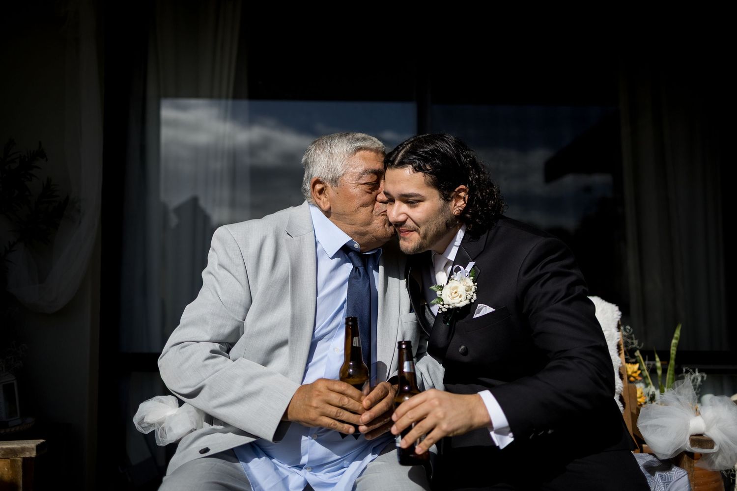 An elderly man in a gray suit kisses a groom in a black tuxedo on the cheek while both hold beer bottles.