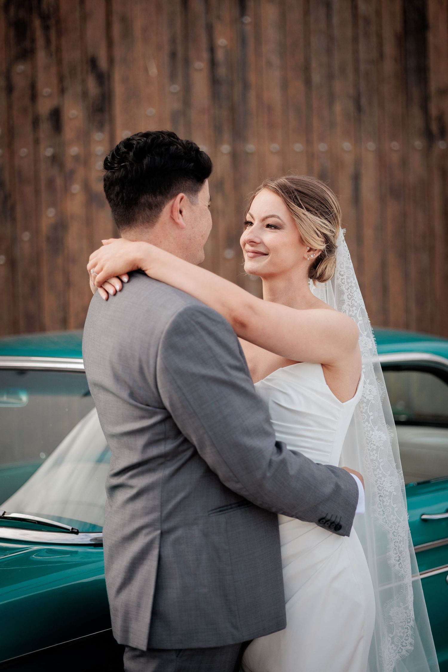 Smiling bride in white gown and veil embracing groom in gray suit by a classic teal car.