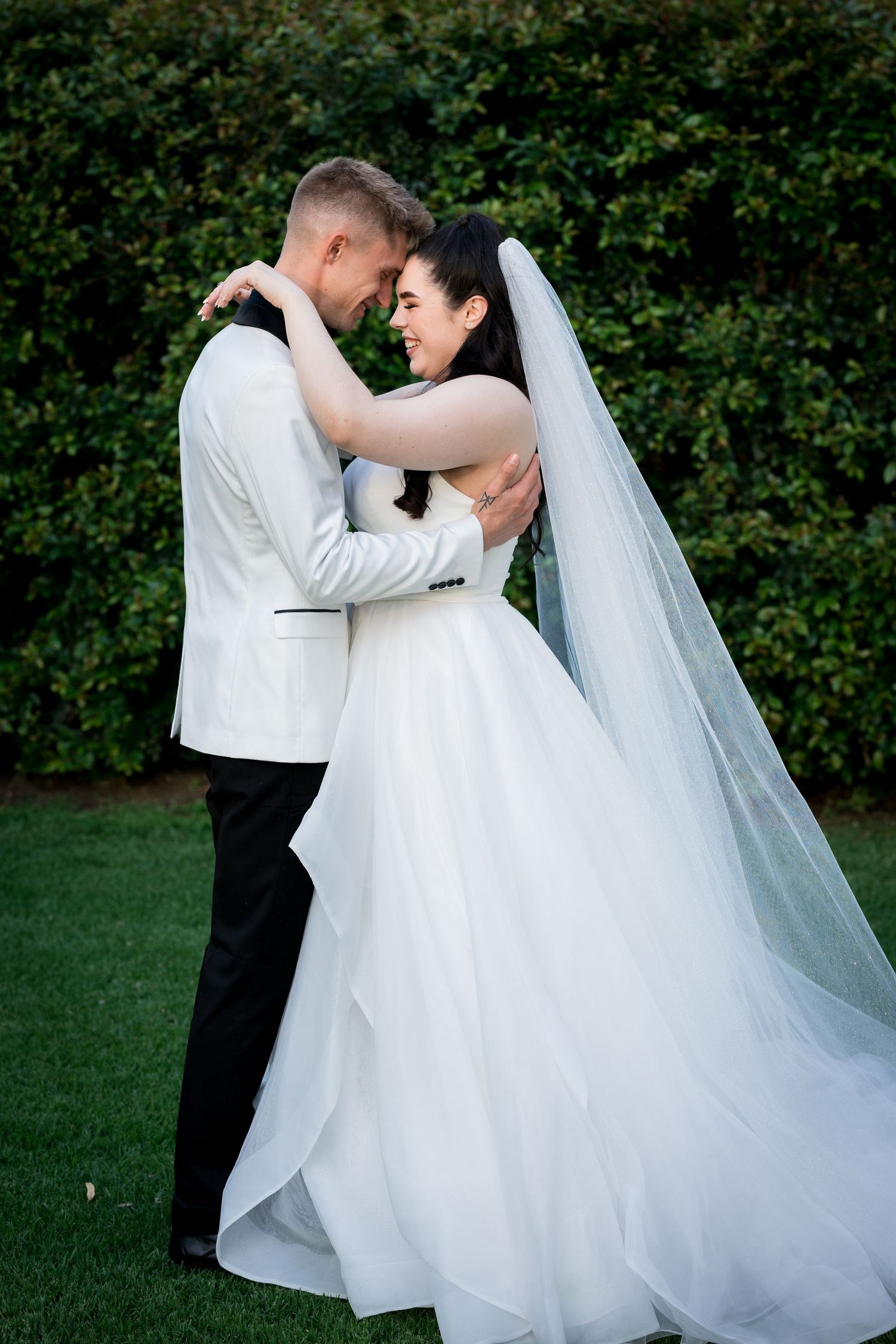 Bride and groom embrace outdoors on green grass, wearing white wedding attire with veil flowing, surrounded by lush hedges.