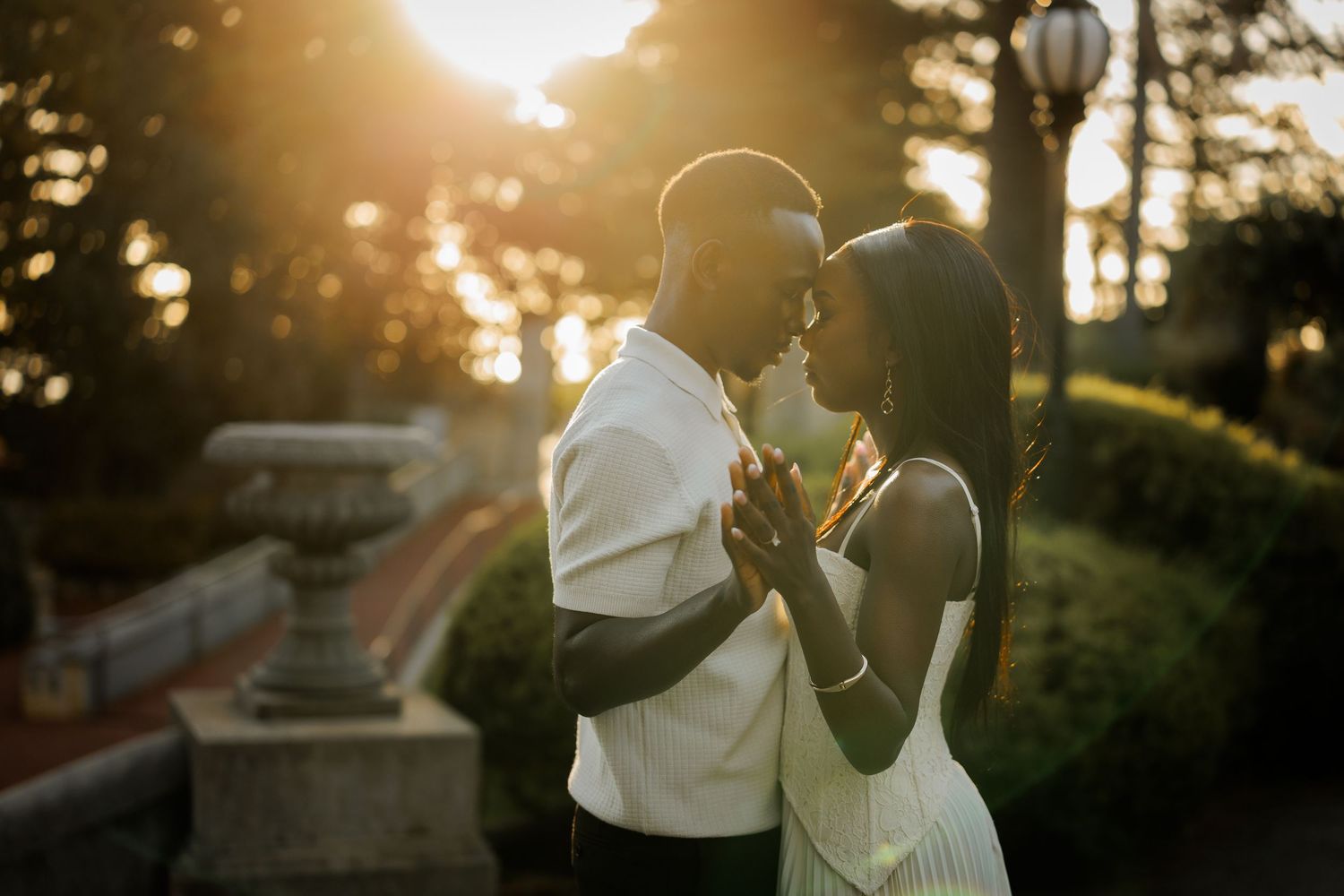 Couple embracing in an intimate moment in a garden at golden hour sunset, with warm sunlight filtering through the trees behind them.