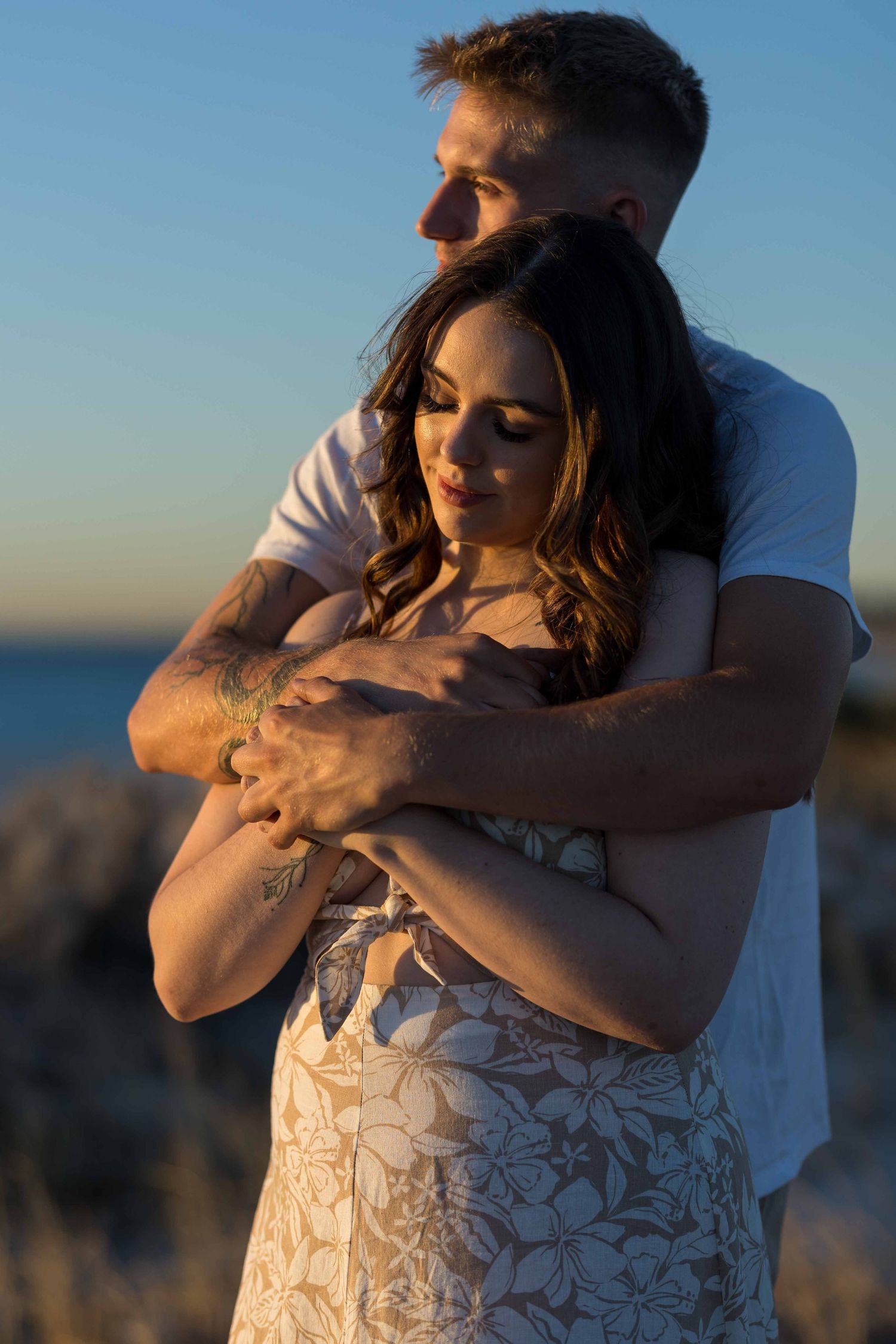 Couple embracing at golden hour sunset by the ocean, man wrapping arms around woman in floral dress.