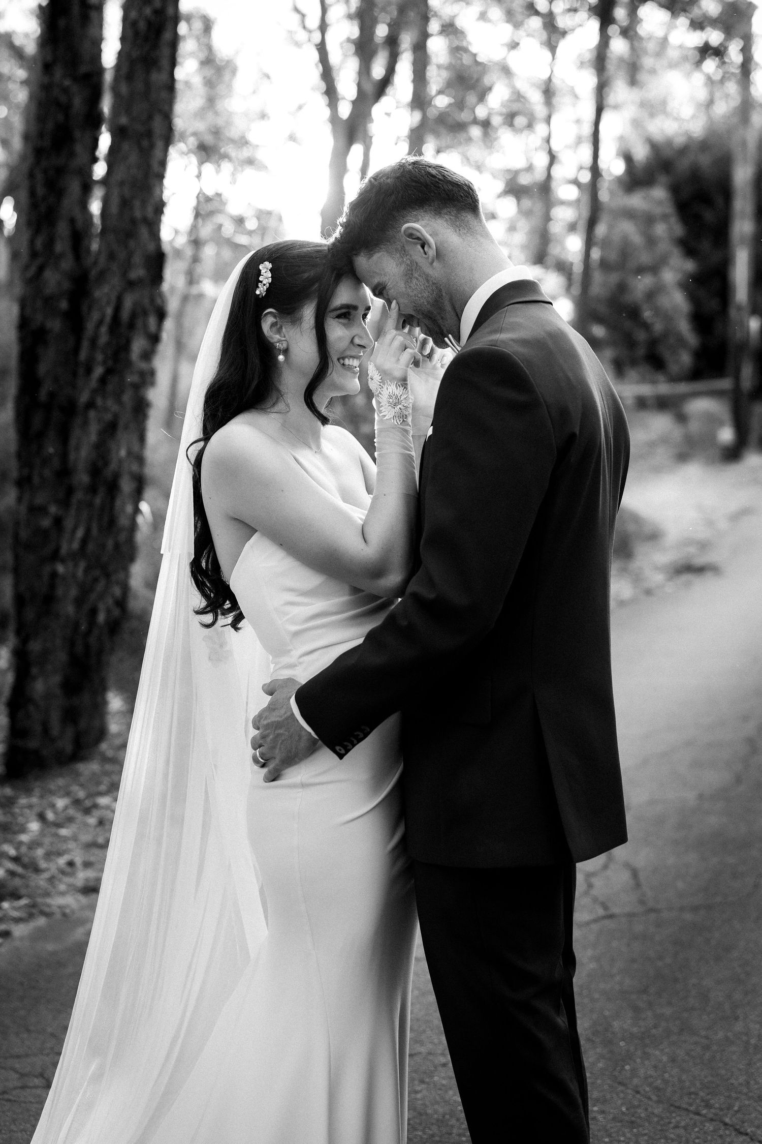 Black and white photo of a smiling bride in a white gown and veil embracing a groom in a suit outdoors.