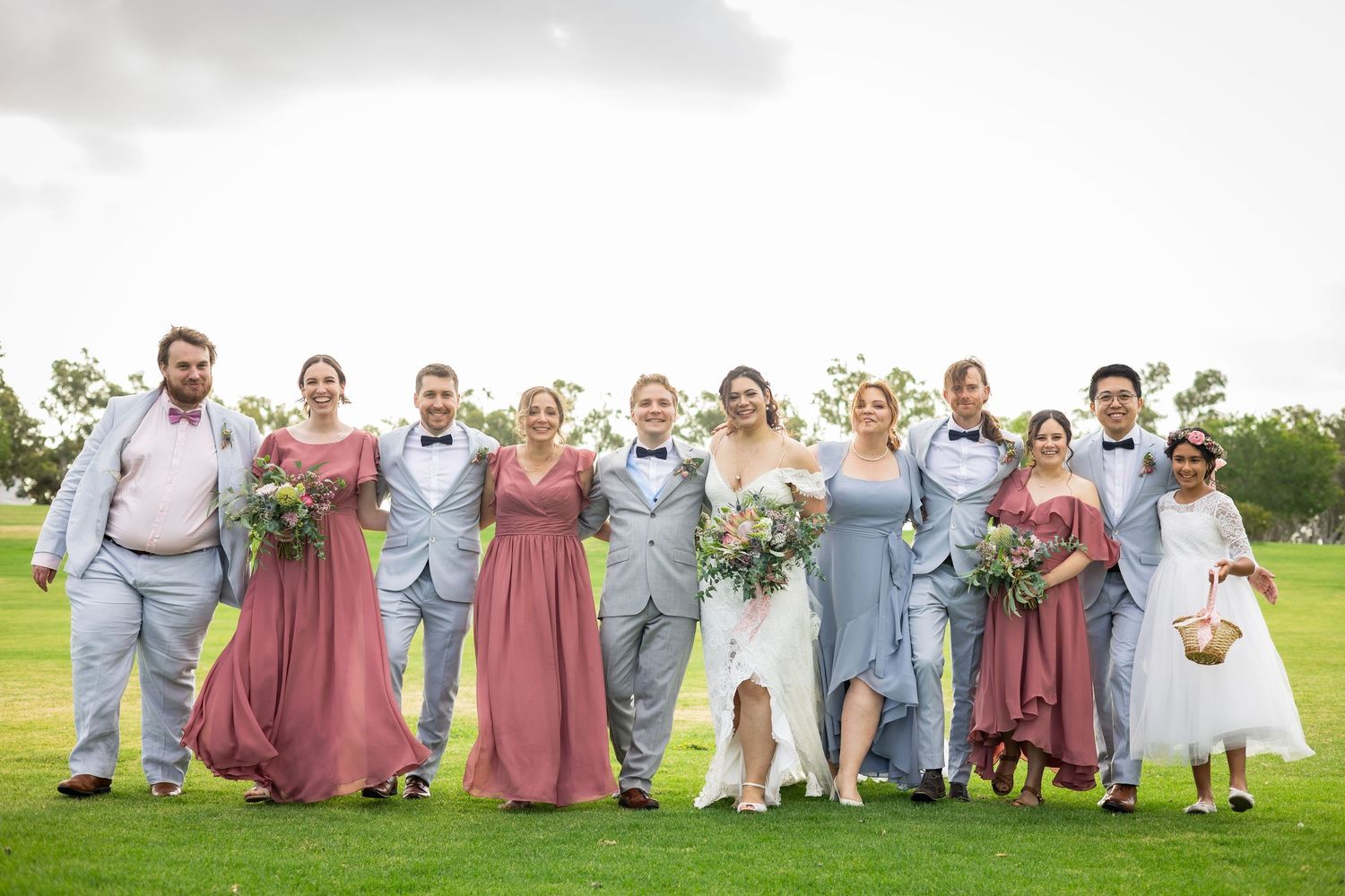 Wedding party walking together on green lawn, bridesmaids in pink and blue gowns with groomsmen in gray suits.