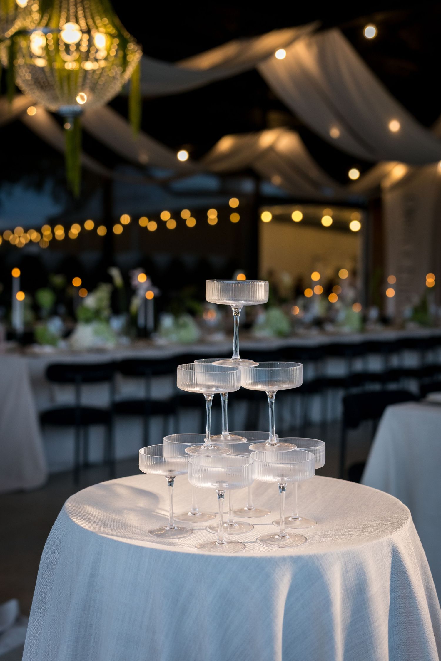 Elegant champagne tower on white draped table at romantic wedding reception with string lights.