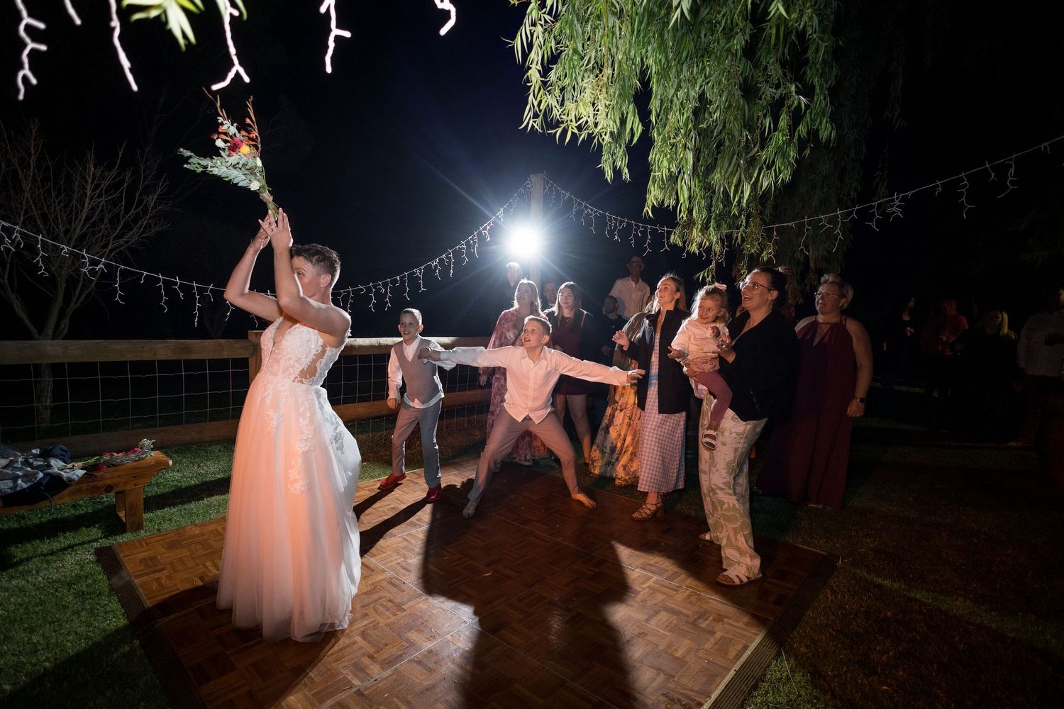 Wedding party dancing under string lights at an outdoor nighttime reception celebration.