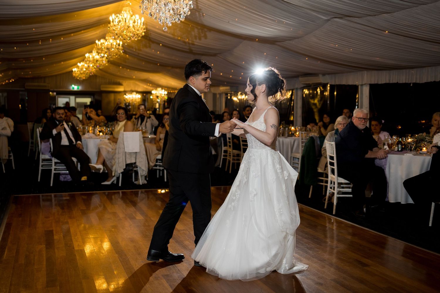 Newlyweds share their first dance on wooden floor under crystal chandeliers.