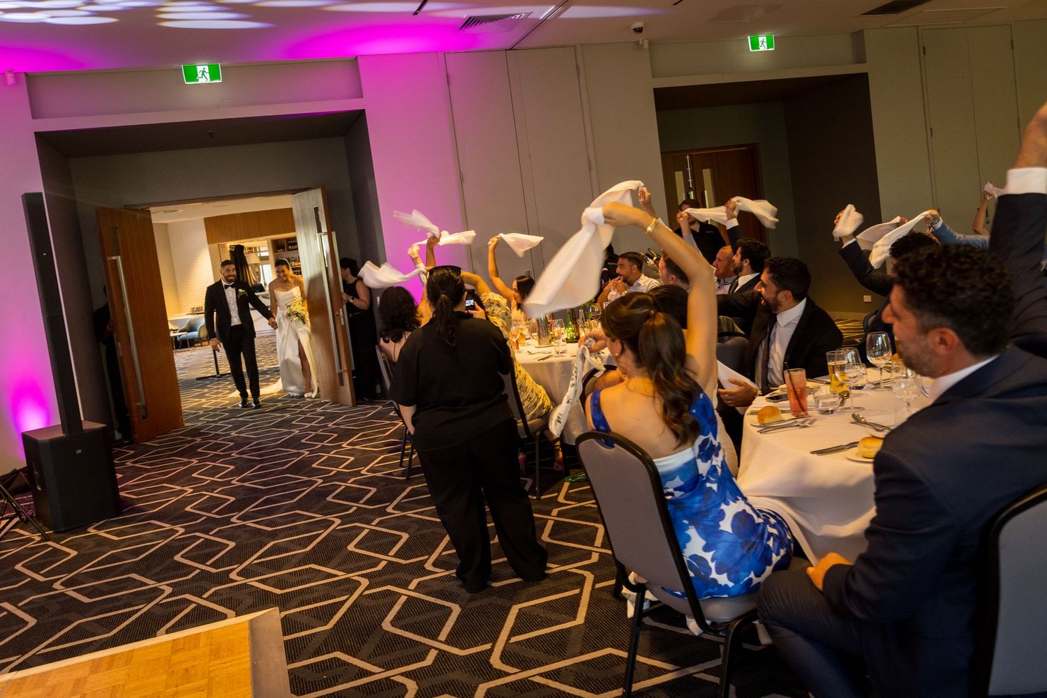 Excited wedding guests wave napkins in the air during a lively reception entrance at an elegant ballroom.