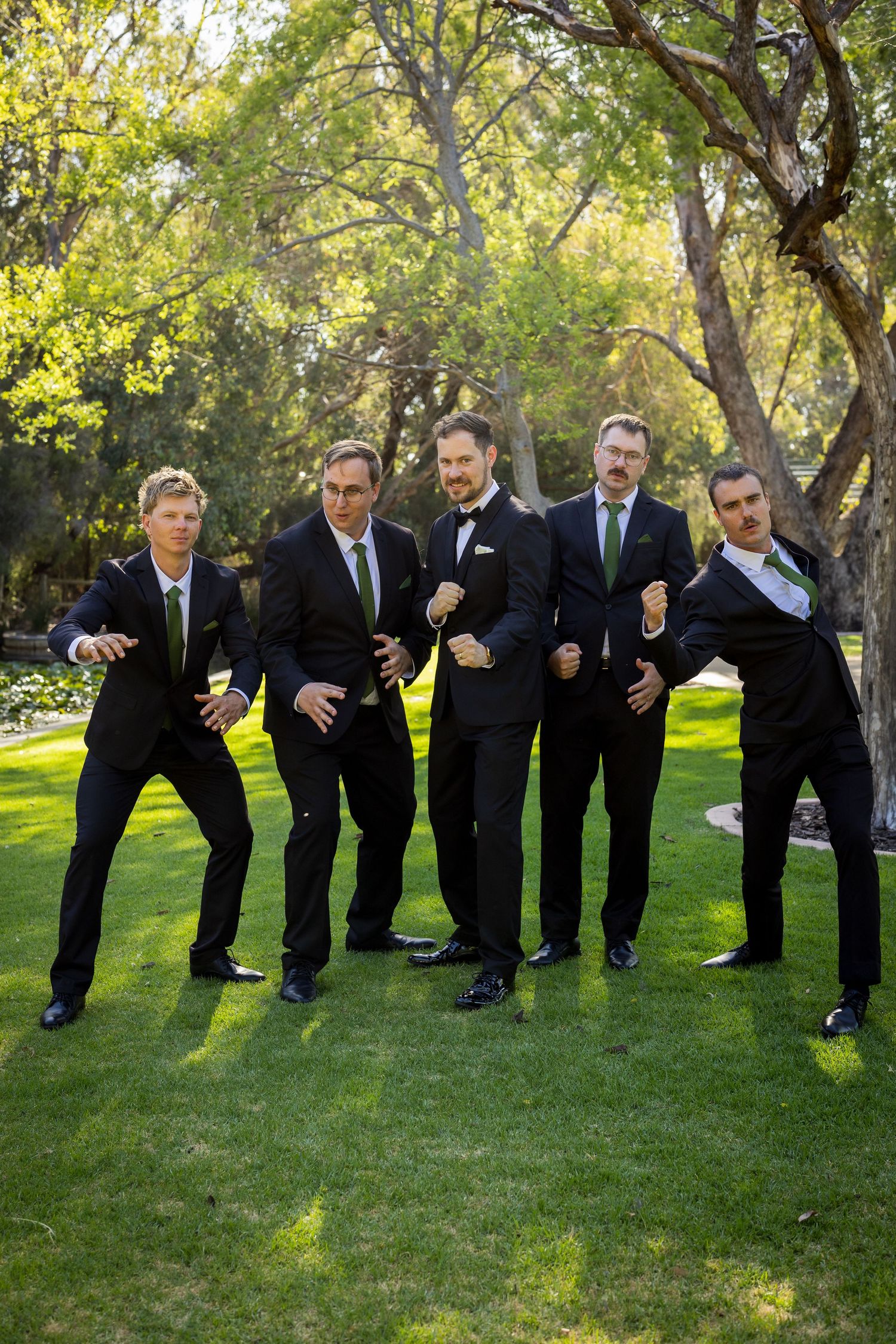 Five men in black suits with green ties pose playfully on grass with trees in background at a wedding.