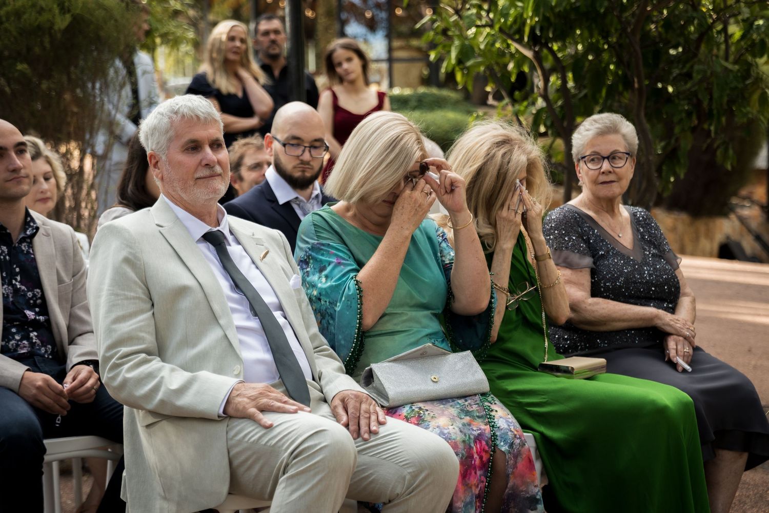 Emotional wedding guests seated outdoors, with a woman in green dress wiping tears during the ceremony.
