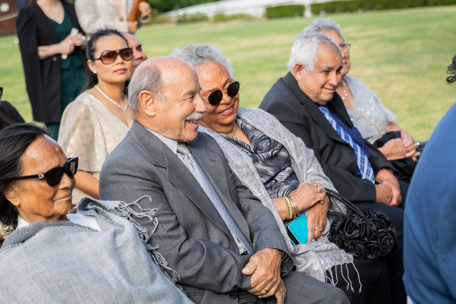 Elderly wedding guests laugh and smile while seated outdoors, wrapped in silver shawls at a wedding ceremony.