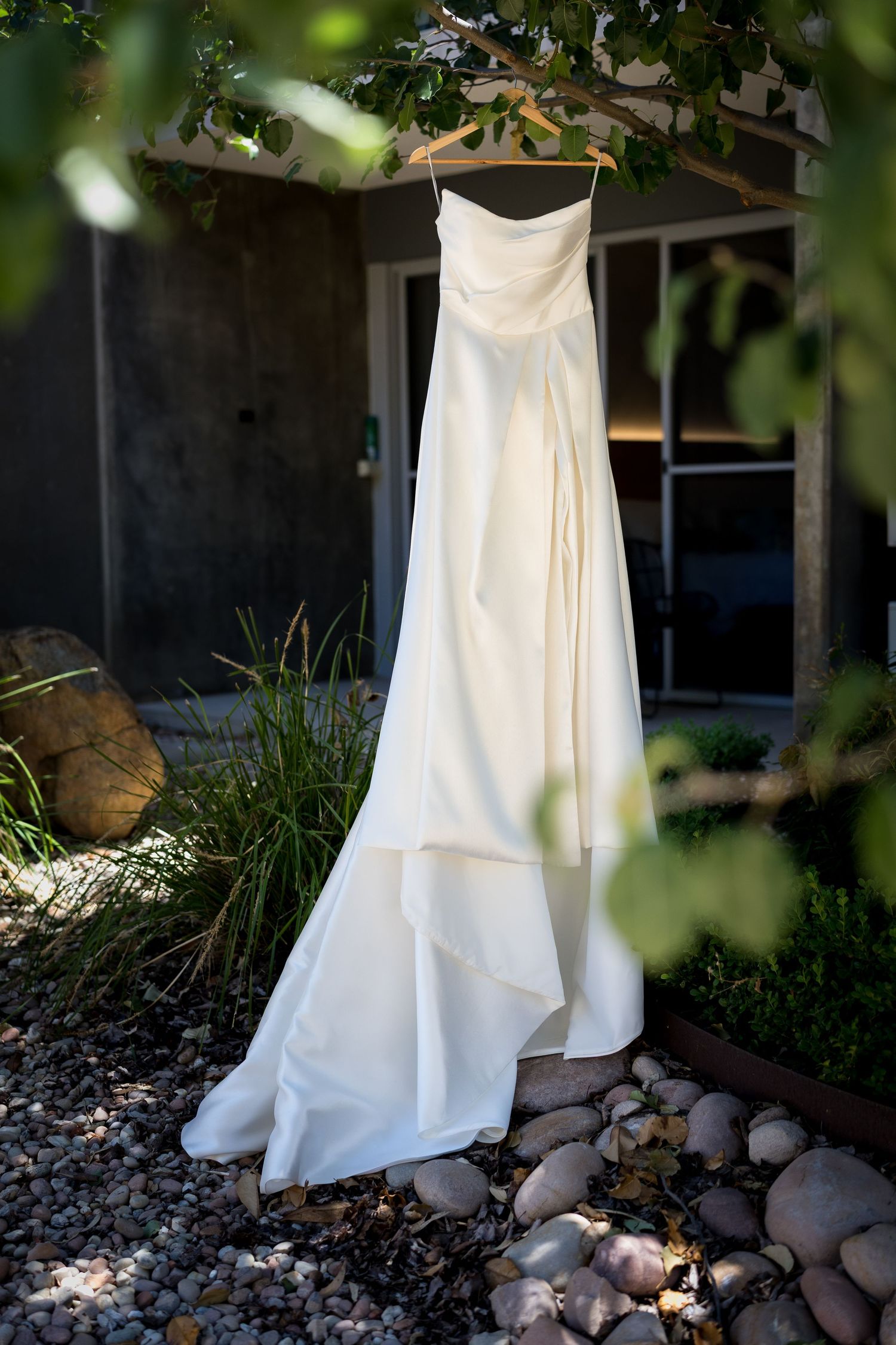 Elegant strapless white wedding dress hanging on a wooden hanger from a tree branch outdoors.