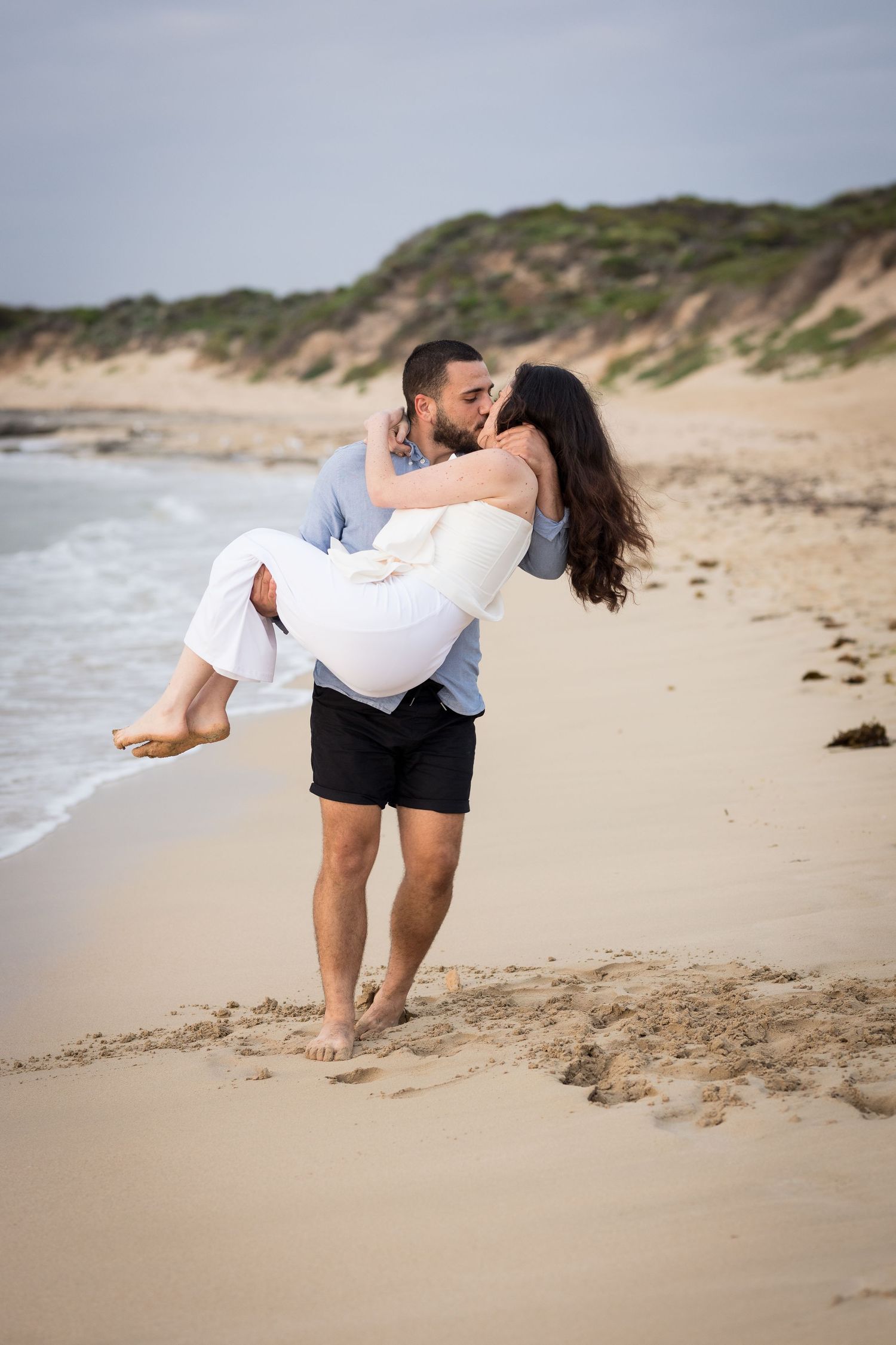 Man carrying woman in white dress sharing kiss on sandy beach.