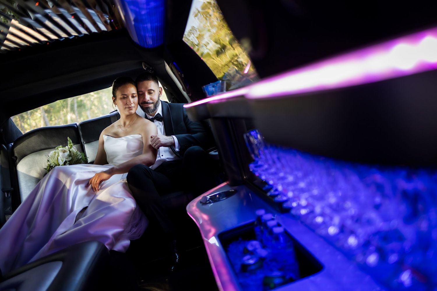 Newlywed couple sitting together in a decorated limousine with purple lights on their wedding day.