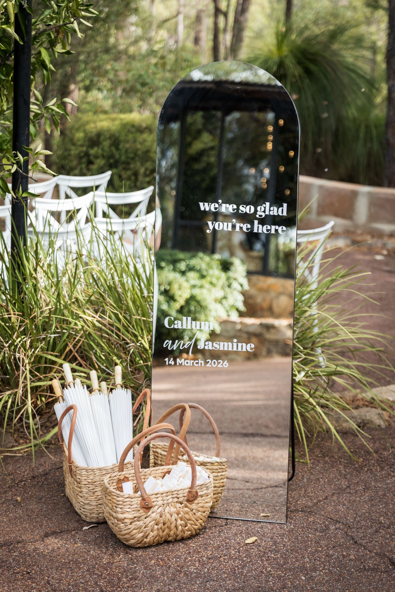 Elegant wedding welcome mirror sign for Callum and Jasmine with wicker baskets and white umbrellas outdoors.