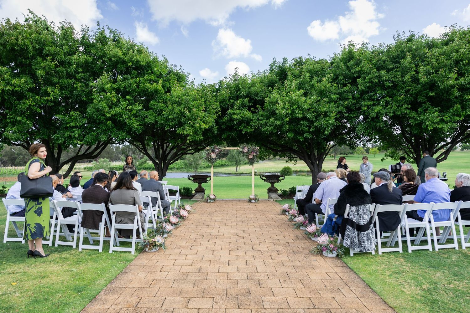 Outdoor wedding ceremony with guests seated on white chairs under lush green trees on a sunny day.