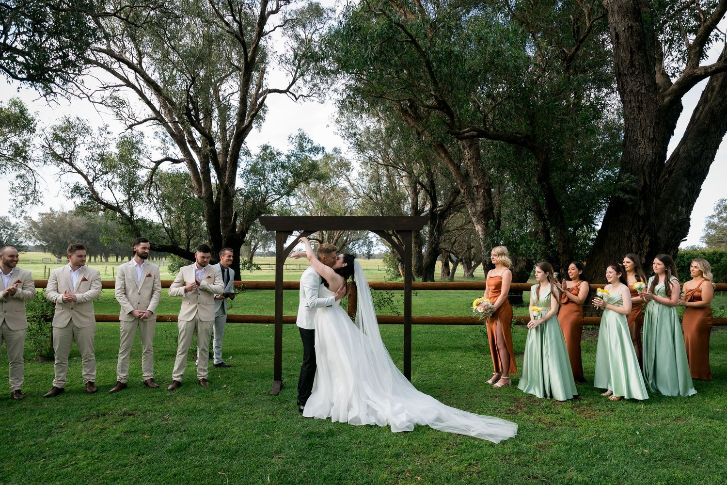 Wedding party lined up outdoors with bridesmaids in pastel dresses and groomsmen in light suits.
