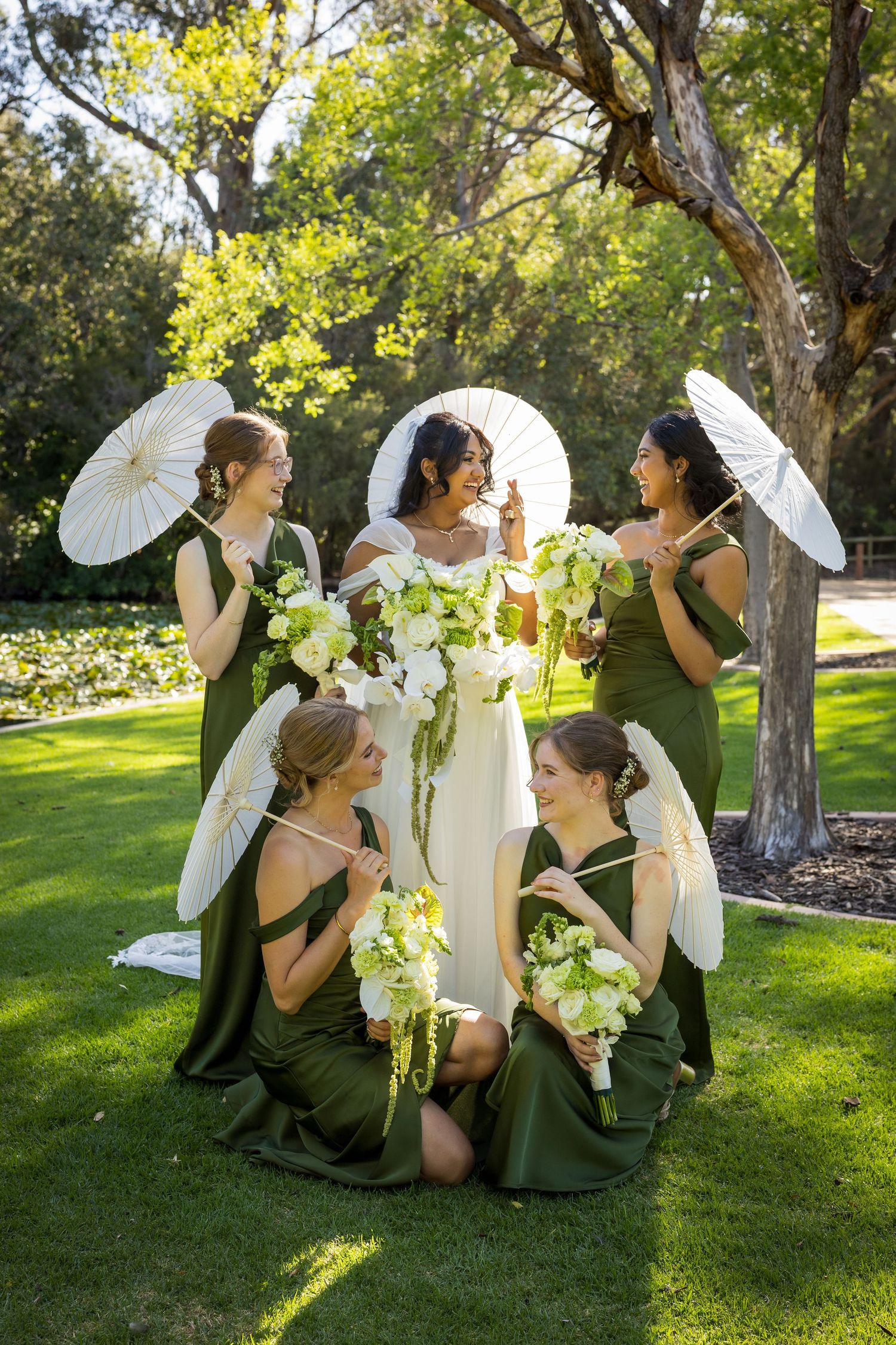 Bride in white gown poses with four bridesmaids in olive green dresses holding parasols and bouquets.