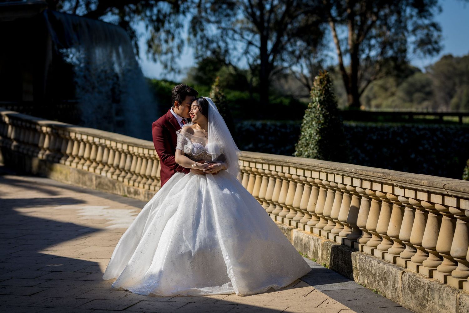 Bride in white ball gown and groom in red suit share a romantic kiss on a stone terrace surrounded by lush gardens.