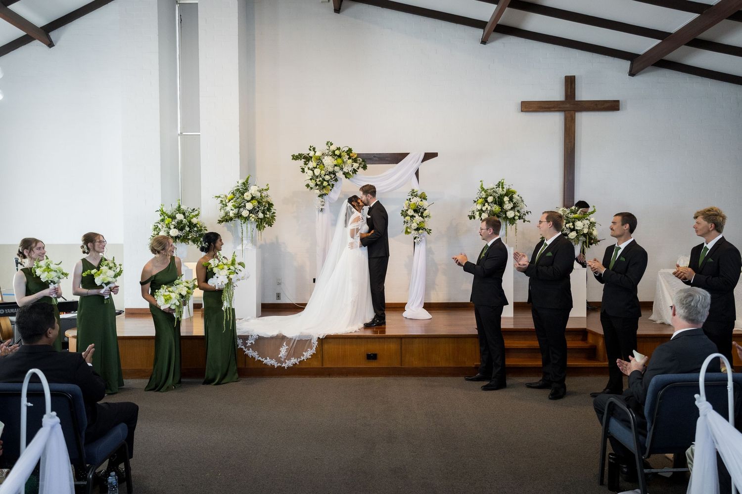 Bride and groom share first kiss at church altar surrounded by wedding party in green dresses and black suits.
