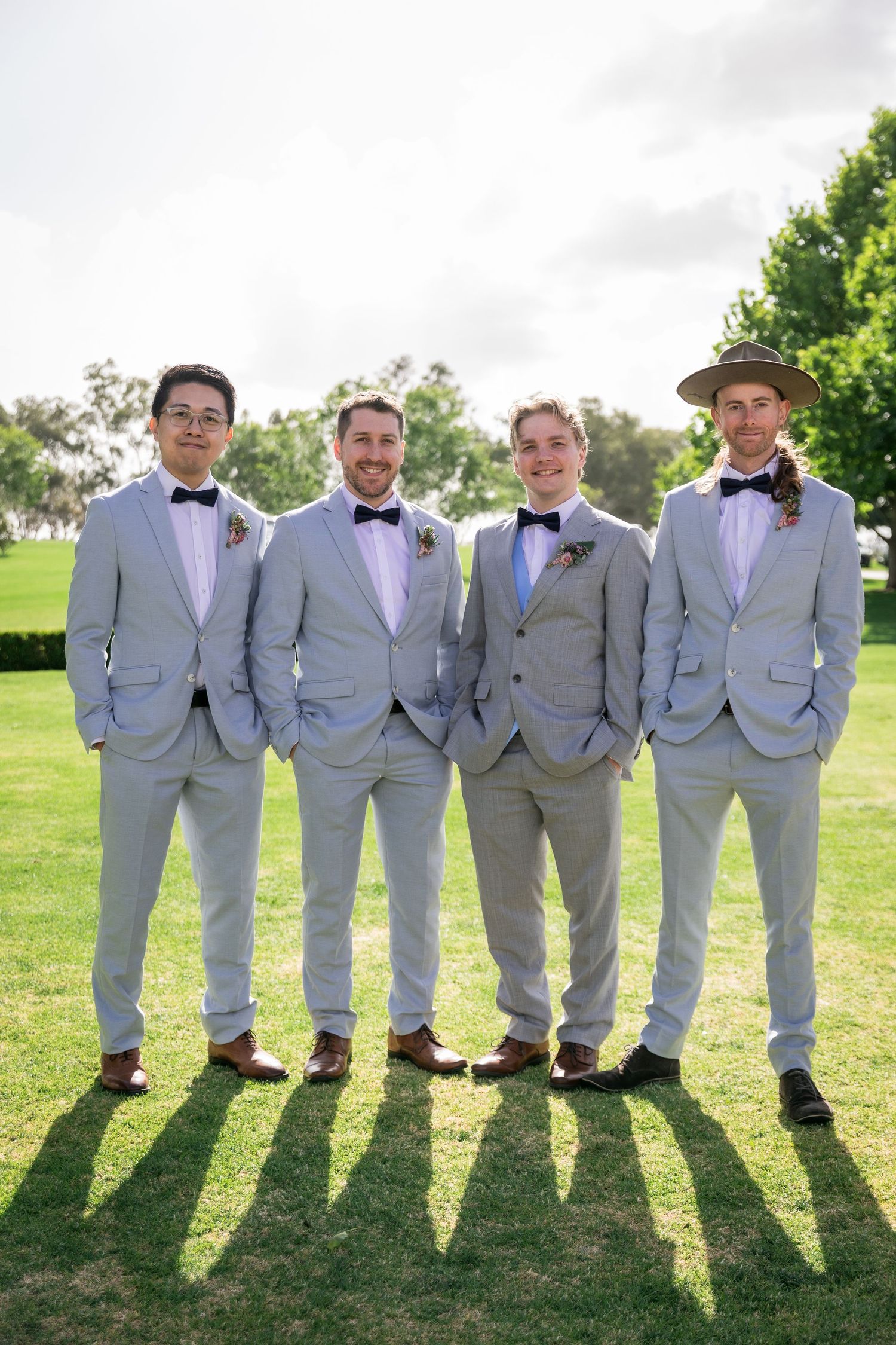 Four groomsmen in light grey suits and bow ties posing on green lawn.