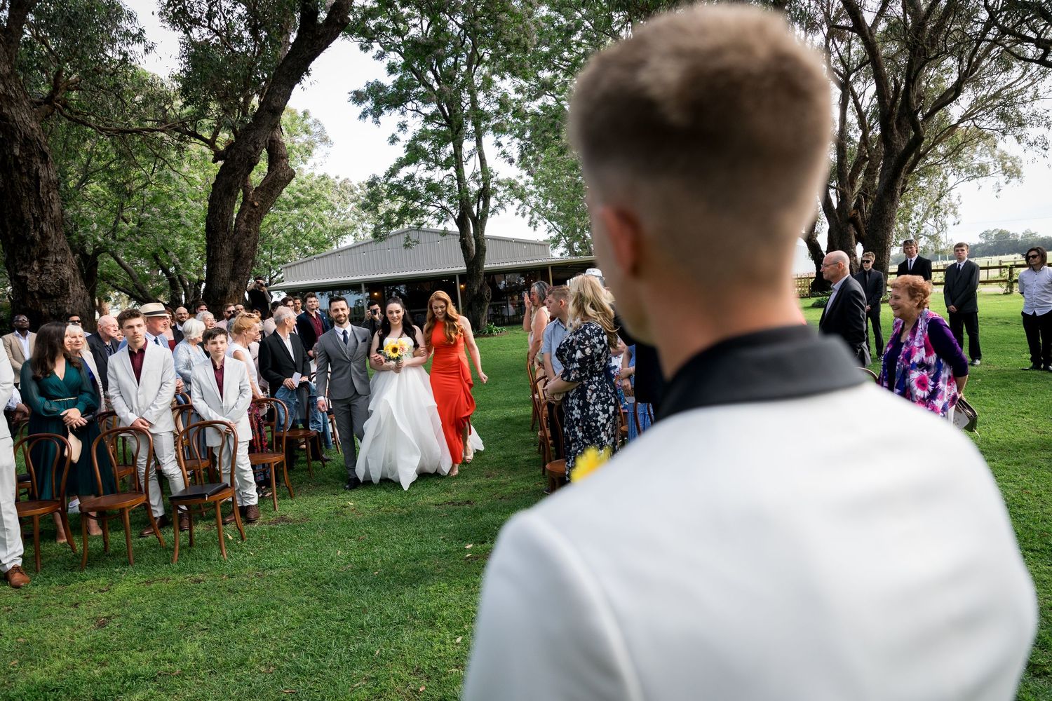 Wedding ceremony guests gathered on a lush green lawn surrounded by trees.
