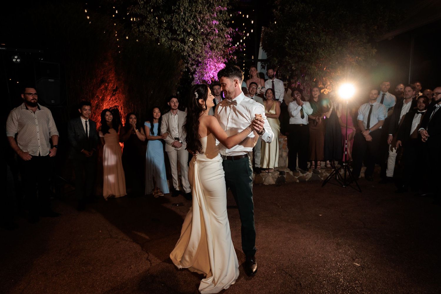 Bride and groom share their first dance outdoors at night, surrounded by wedding guests under warm string lights.