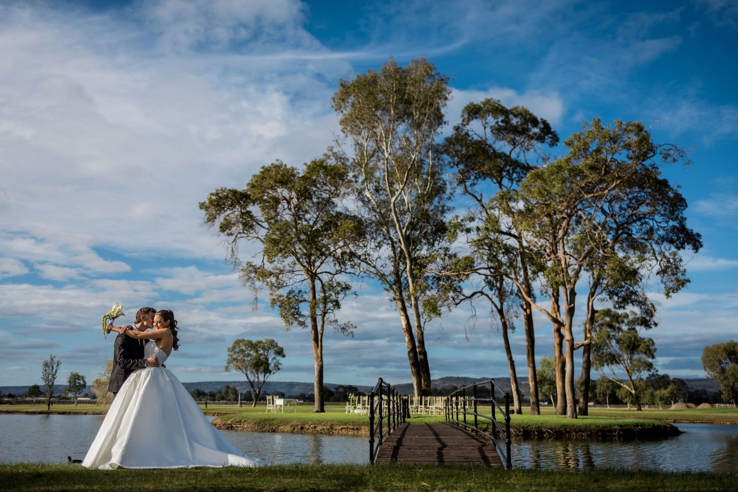 Bride and groom share a romantic kiss by a serene lake surrounded by tall trees under a vibrant blue sky.