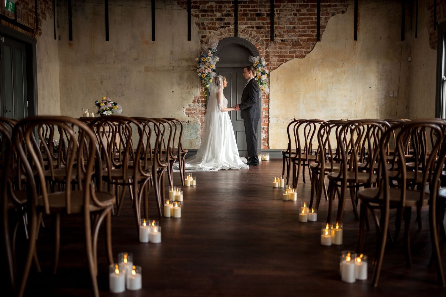 Romantic wedding ceremony in rustic venue with candlelit aisle, bentwood chairs, and couple standing beneath an arched doorway.