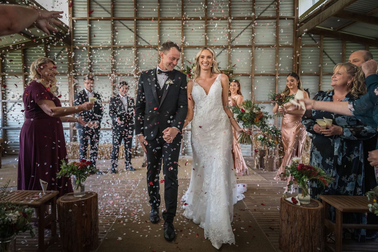 Bride and groom walk down aisle as guests throw confetti in rustic barn wedding venue.