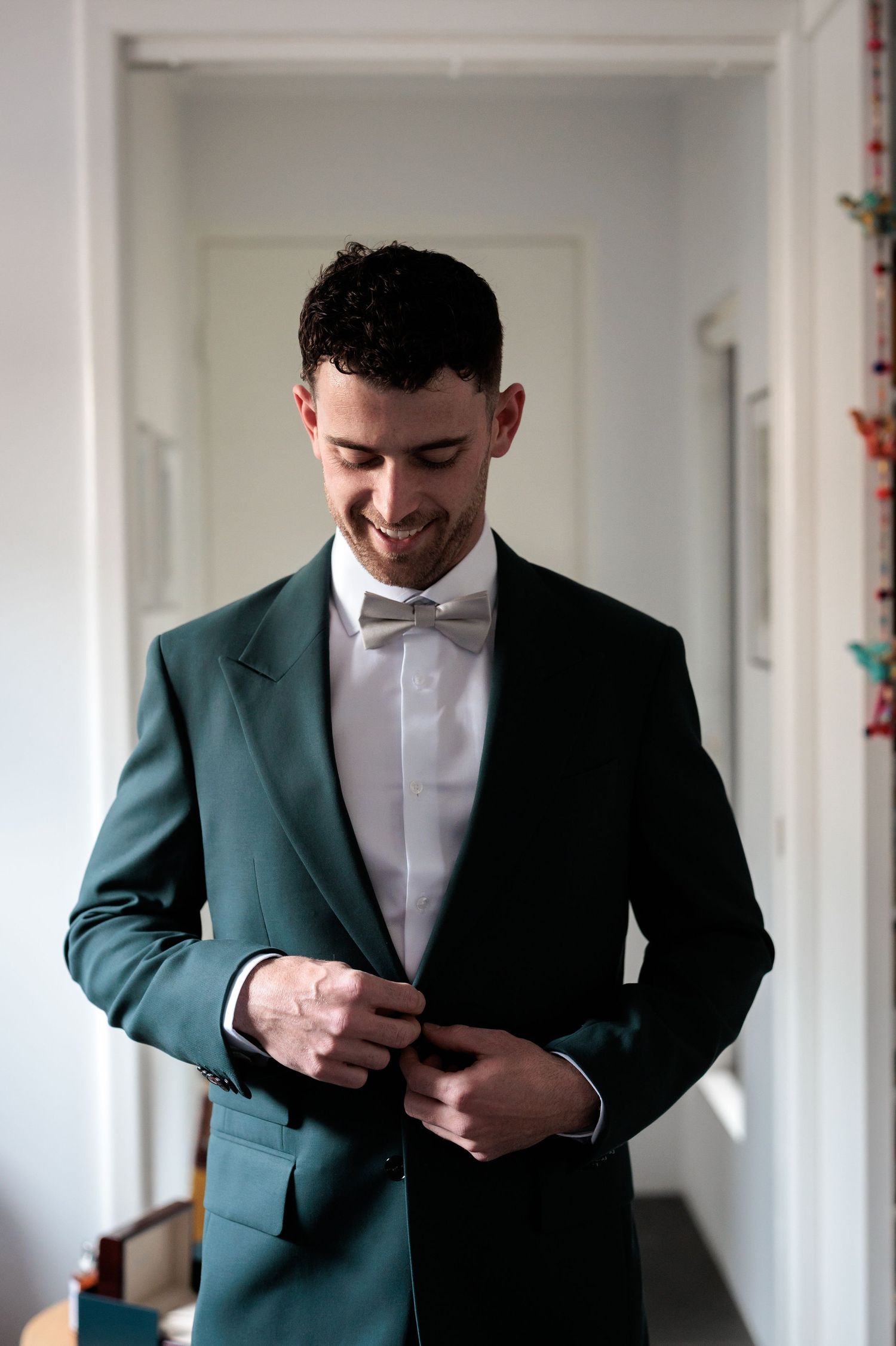 Smiling groom in dark green tuxedo jacket and white bow tie buttoning his jacket while getting ready.