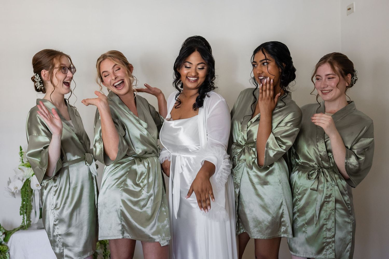 Bride in white robe with four bridesmaids in sage green satin robes laughing and posing together before the wedding.