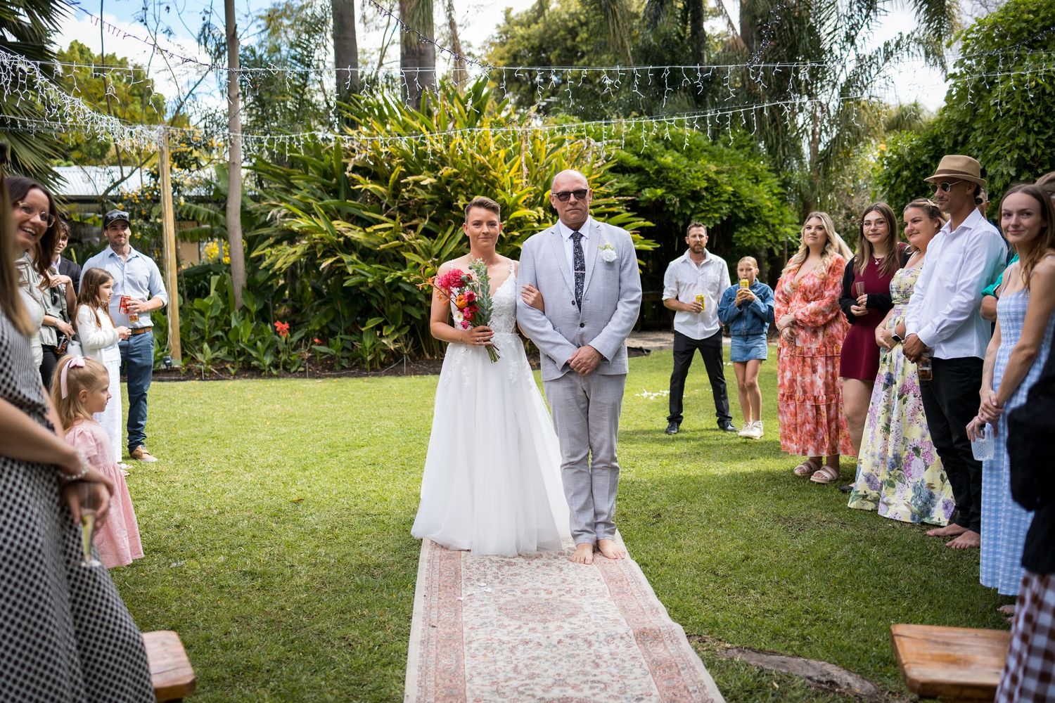 Guests line a garden pathway during an outdoor wedding ceremony.