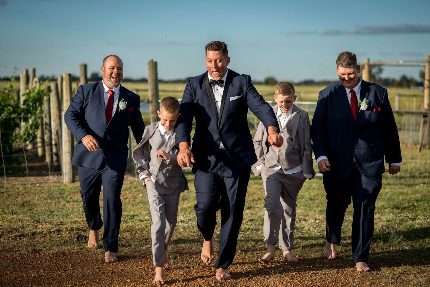 Groomsmen run playfully through field in coordinated navy suits and gray pants.