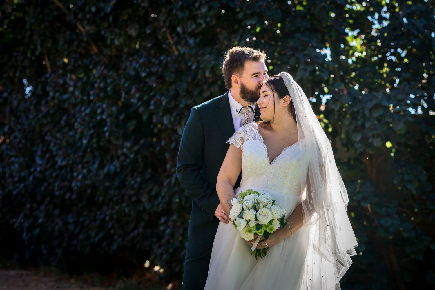 A bride and groom share a romantic moment outdoors, with the bride holding a white bouquet and wearing a veil.