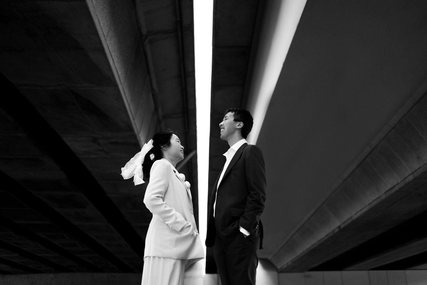 Black and white photo of a couple in formal attire standing under a bridge, gazing upward at dramatic light beams.