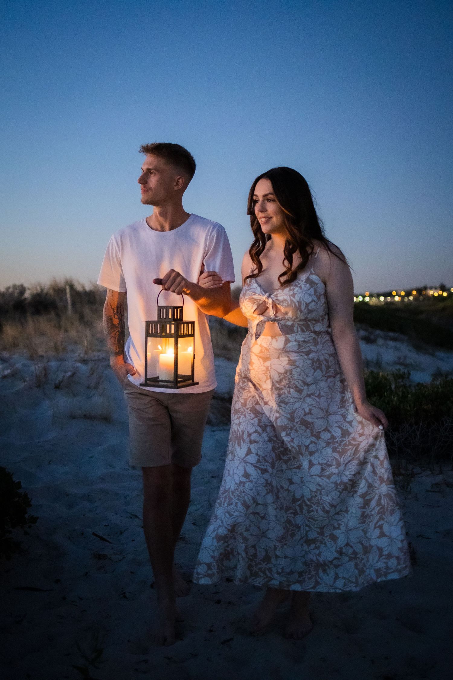 Couple walking on beach at dusk, man holding glowing lantern, woman in floral dress, twilight sky backdrop.