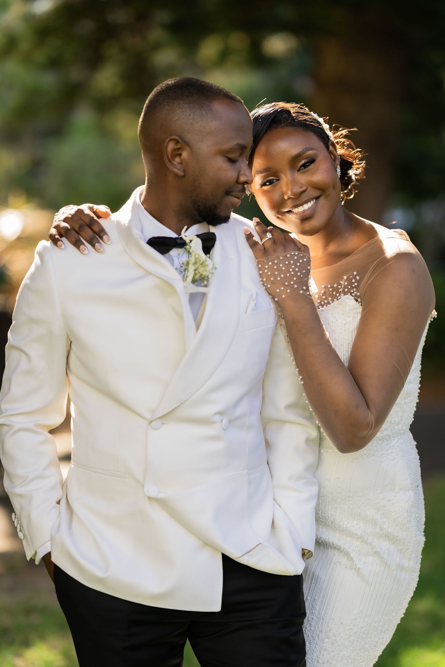 A joyful Black bride and groom in white wedding attire embrace outdoors, smiling warmly in soft golden sunlight.
