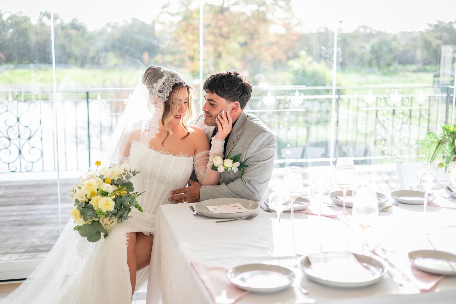 Bride and groom share a romantic moment at elegantly set wedding reception table, bride holding yellow and white bouquet.
