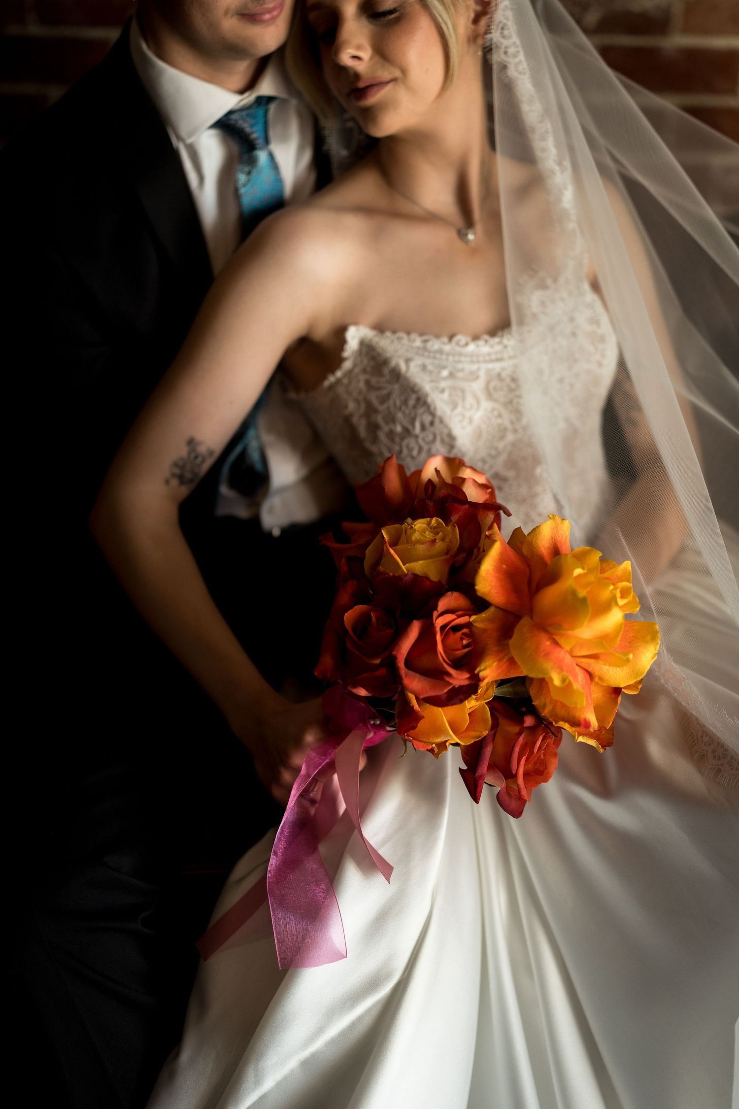 Bride in white lace gown holding vibrant orange and red bouquet with pink ribbon, embraced by groom in black suit.