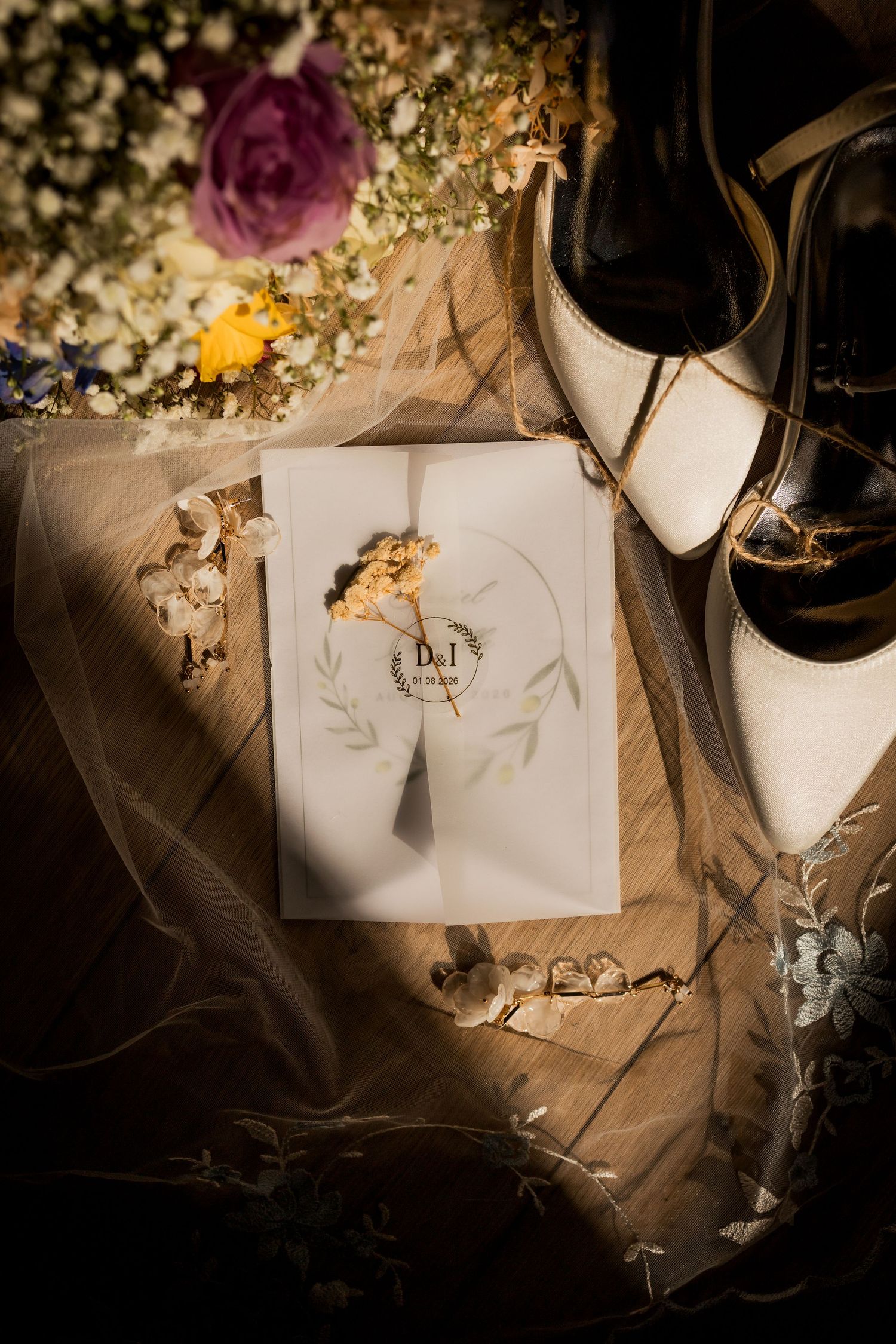 Wedding flat lay with invitation, white heels, floral bouquet, and embroidered veil in golden light.