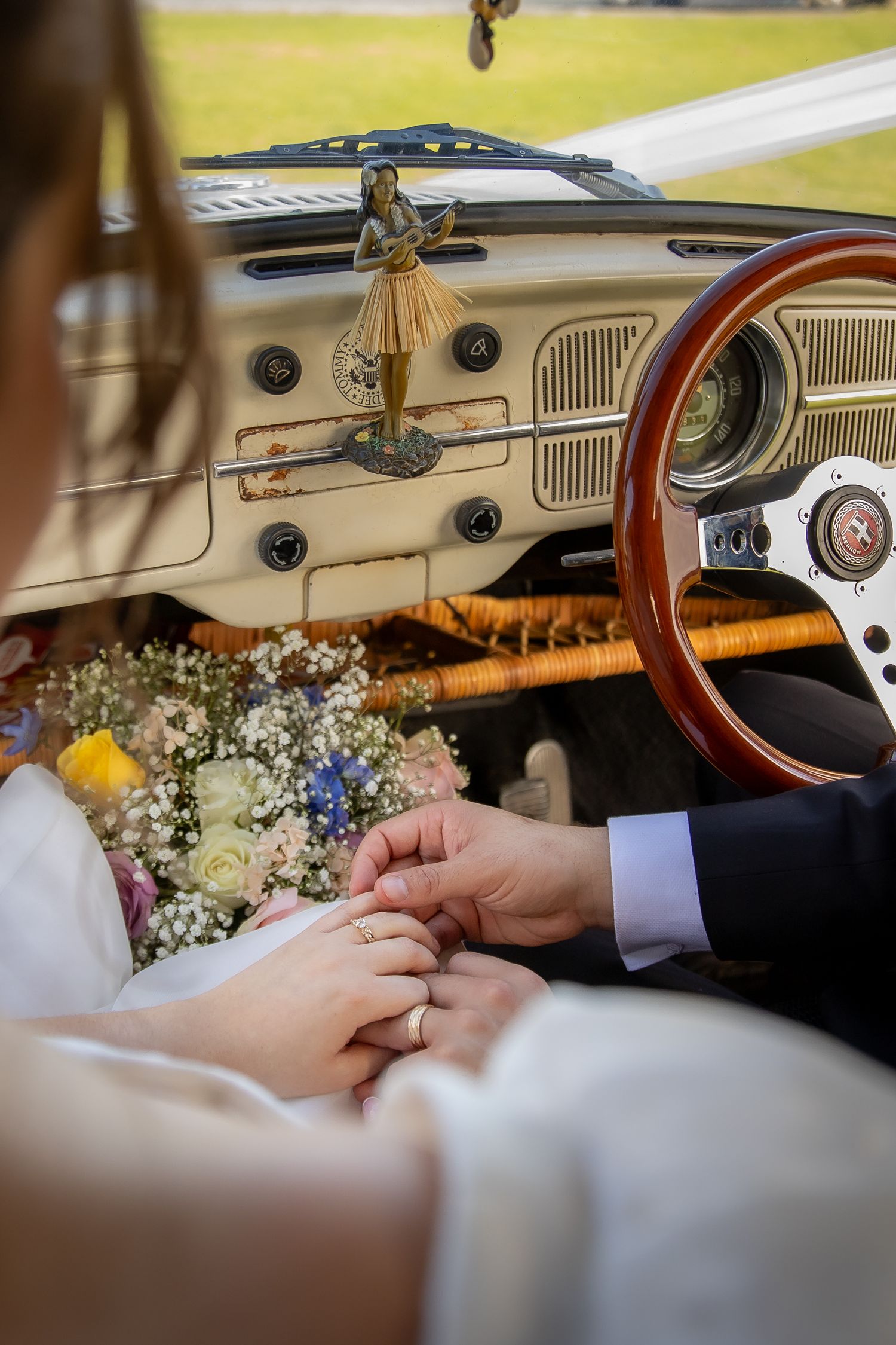 Newlyweds hold hands in a vintage car with a bridal bouquet and hula dancer dashboard ornament.